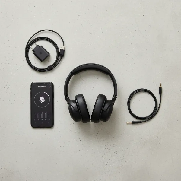 A flat lay of Skullcandy Crusher Evo headphones alongside a smartphone displaying the Skullcandy app interface, a USB-C cable, and a 3.5mm audio jack, highlighting connectivity and app features.
