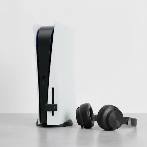 A PS5 console with a USB Bluetooth adapter plugged into its front port, and a pair of generic Bluetooth over-ear headphones resting nearby.